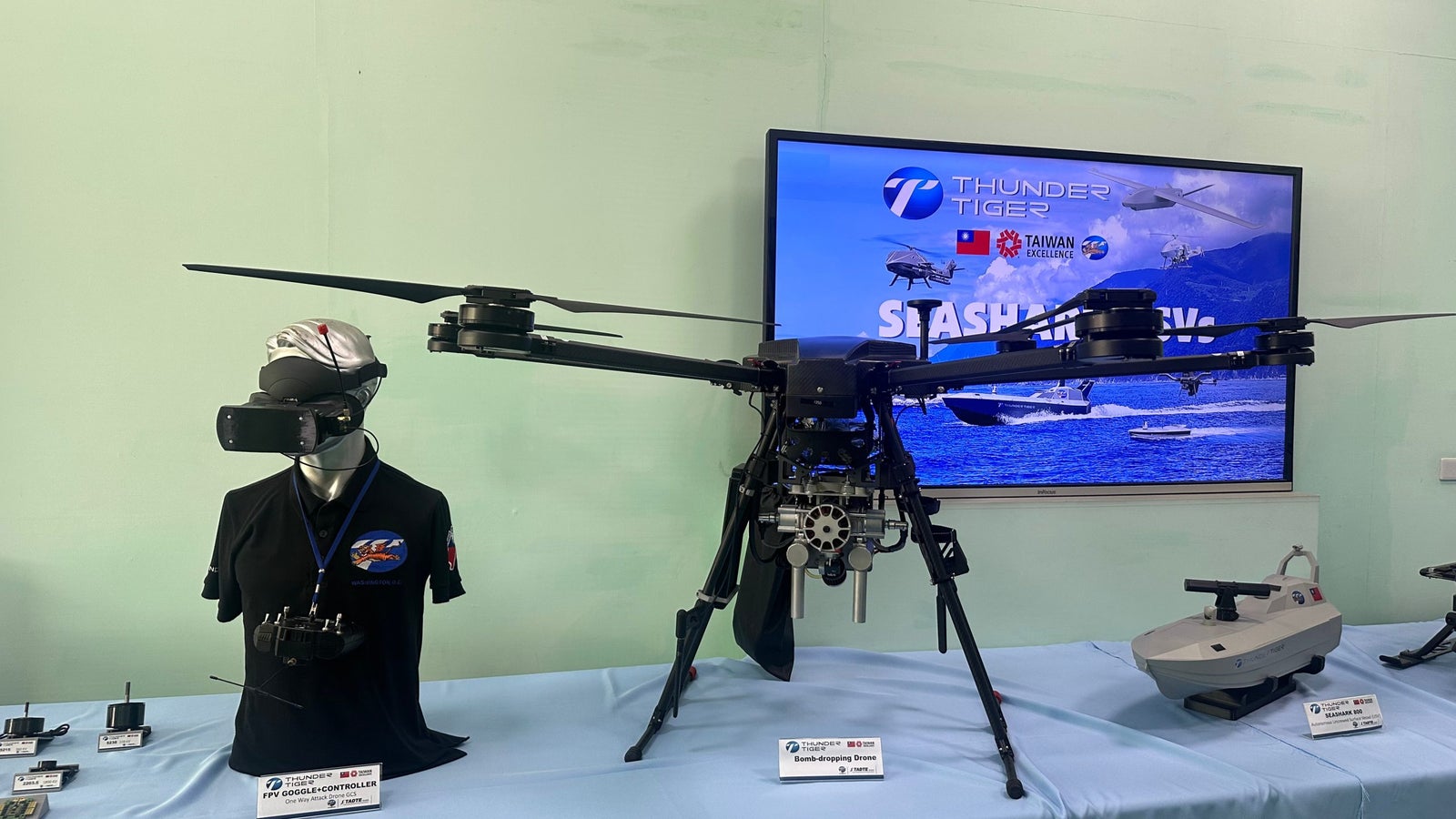 A drone displayed on a table beside a mannequin wearing virtual reality goggles, with a promotional screen in the background.