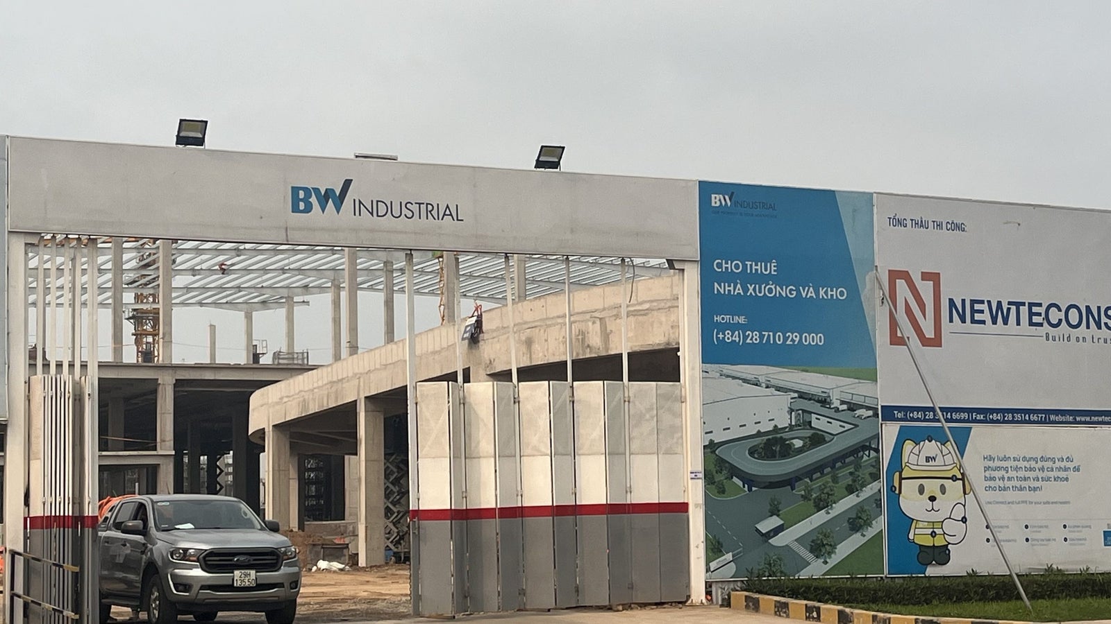 Entrance to a construction site featuring a large sign for BW Industrial and Newtecons, with a silver vehicle parked nearby and building structures in the background.