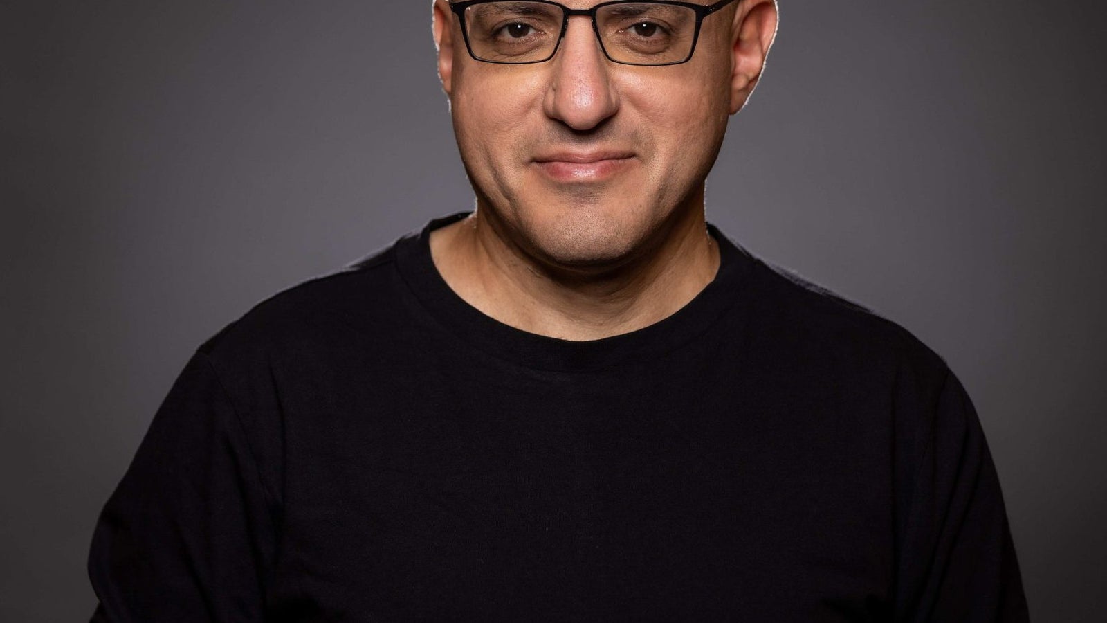 A bald man with glasses wearing a black long-sleeve shirt poses for a portrait against a dark background, smiling slightly.