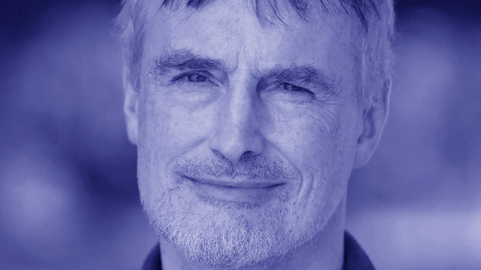 A portrait of a middle-aged man with light hair and a beard, smiling softly, set against a softly blurred background. The image has a blue hue.