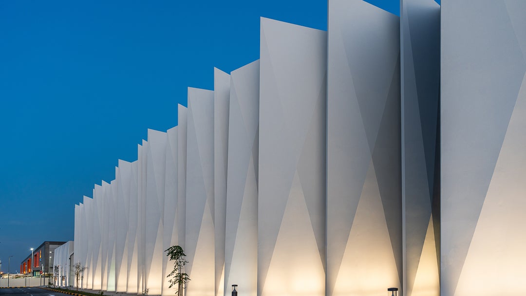 A series of tall, angular, white architectural panels arranged in a row, illuminated at their bases, against a twilight blue sky, with a small tree and light poles visible along a paved road in the foreground.