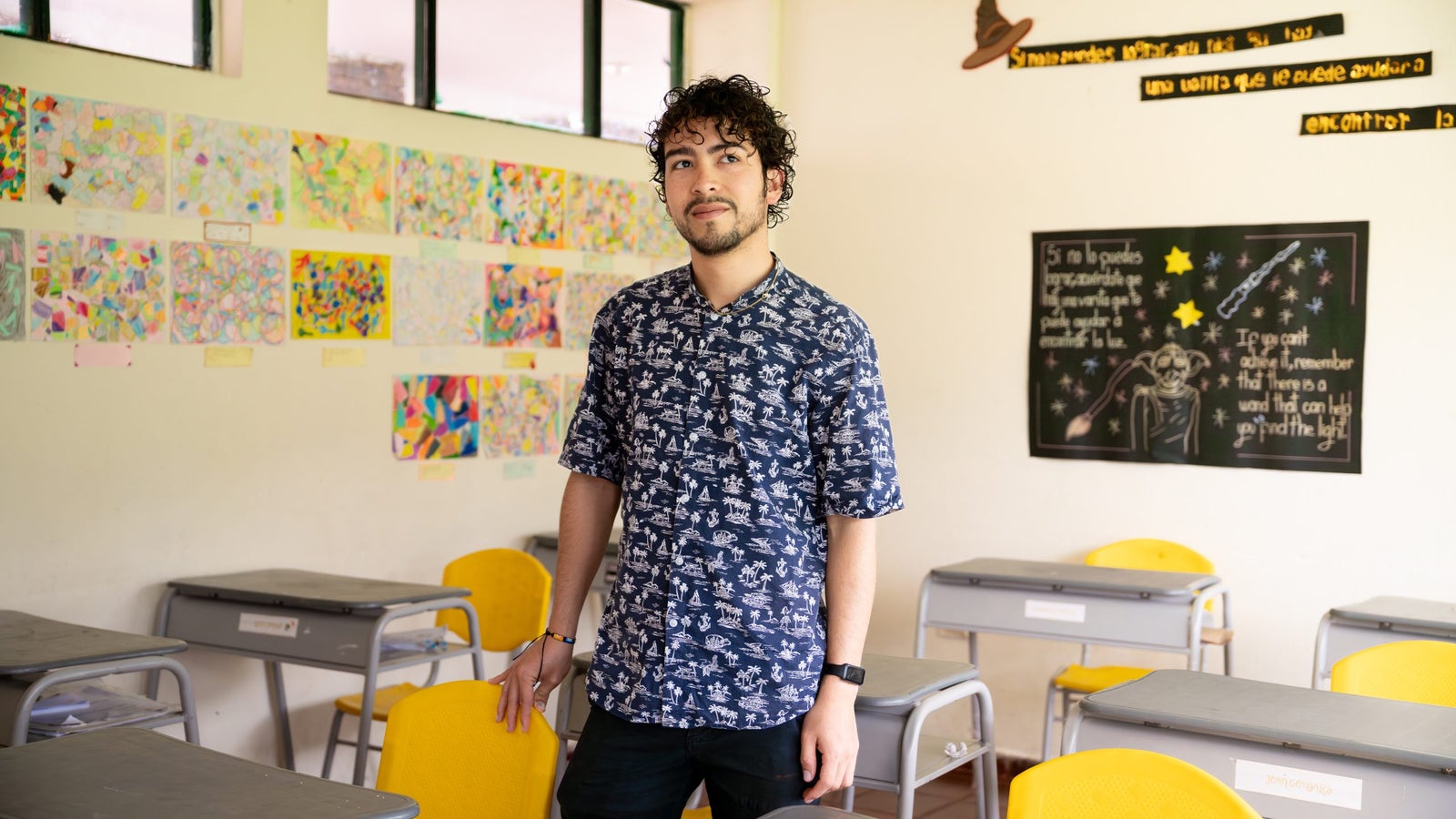 A young man with curly hair stands in a brightly lit classroom. He wears a blue patterned shirt and is positioned next to a yellow chair. Colorful student artwork is displayed on the walls, and there is a chalkboard with inspirational messages in the background.