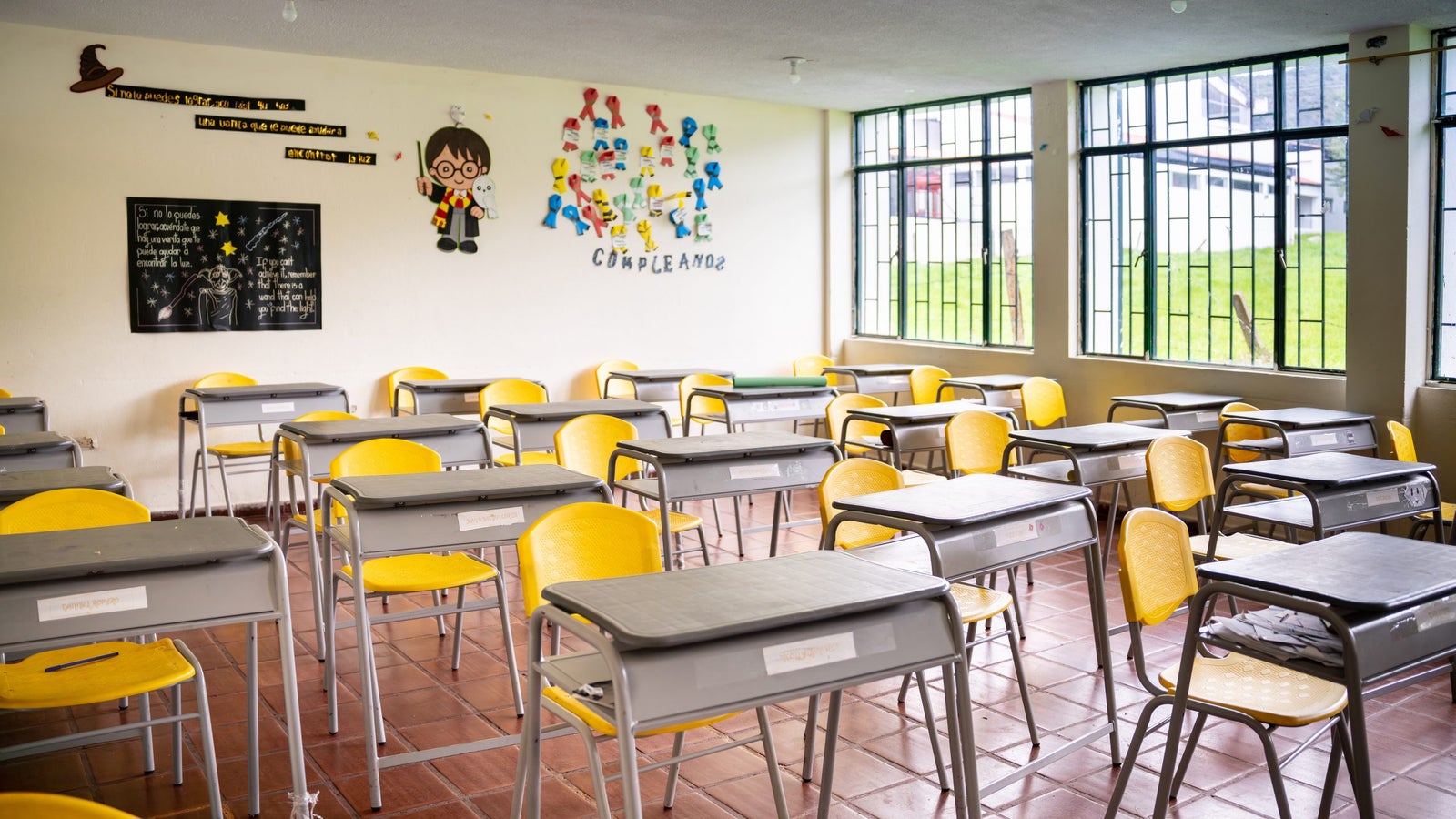 A brightly lit classroom featuring several yellow chairs and gray desks arranged in rows, with educational decorations on the walls, including cartoon characters and motivational quotes written in Spanish.