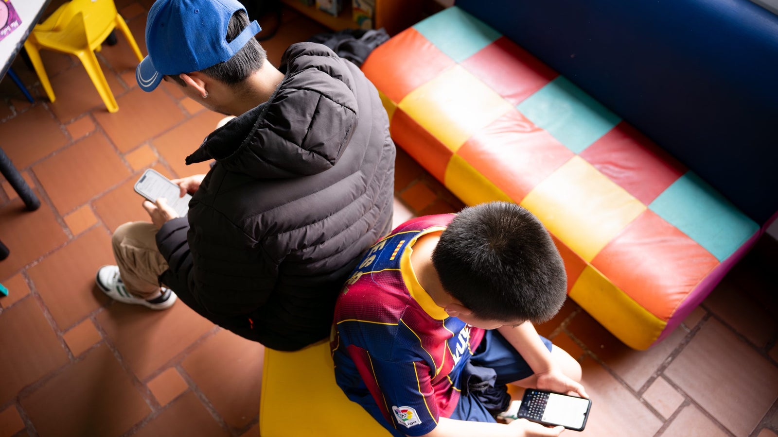 A young man in a black jacket and blue cap sits on a yellow bench, using a smartphone, while a boy in a red and blue jersey sits nearby on a colorful checkered bench, using a small handheld device. The setting appears to be a cozy indoor space with tiled flooring and bookshelves in the background.