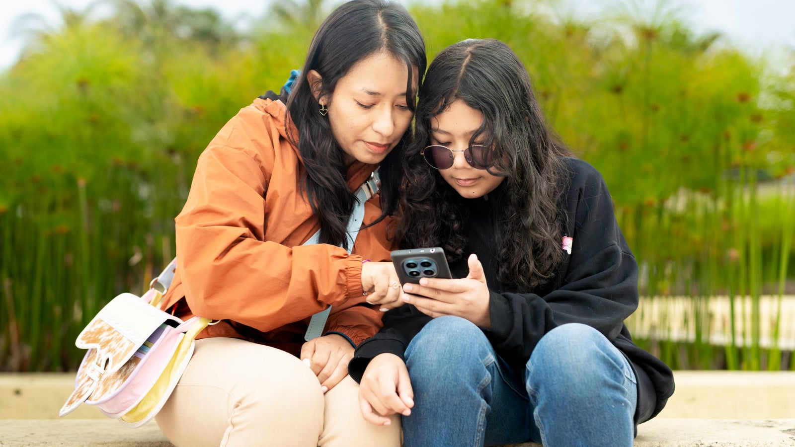 Two young women sitting closely together on a concrete bench, closely examining a smartphone. One woman is wearing an orange jacket and has long dark hair, while the other is wearing sunglasses and a black sweatshirt. Lush greenery is visible in the background.