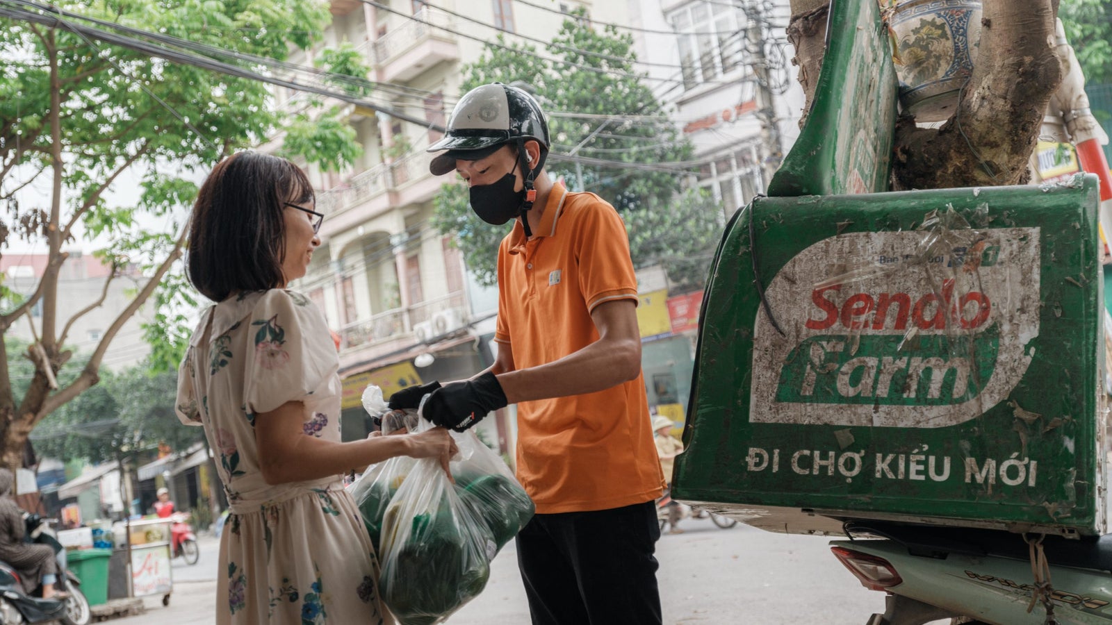 A delivery person in an orange shirt and black mask hands a bag of groceries to a woman wearing a floral dress with short hair. They are on a street with trees and buildings in the background, and a green delivery vehicle with a logo reading 'Sendo Farm' is parked nearby.