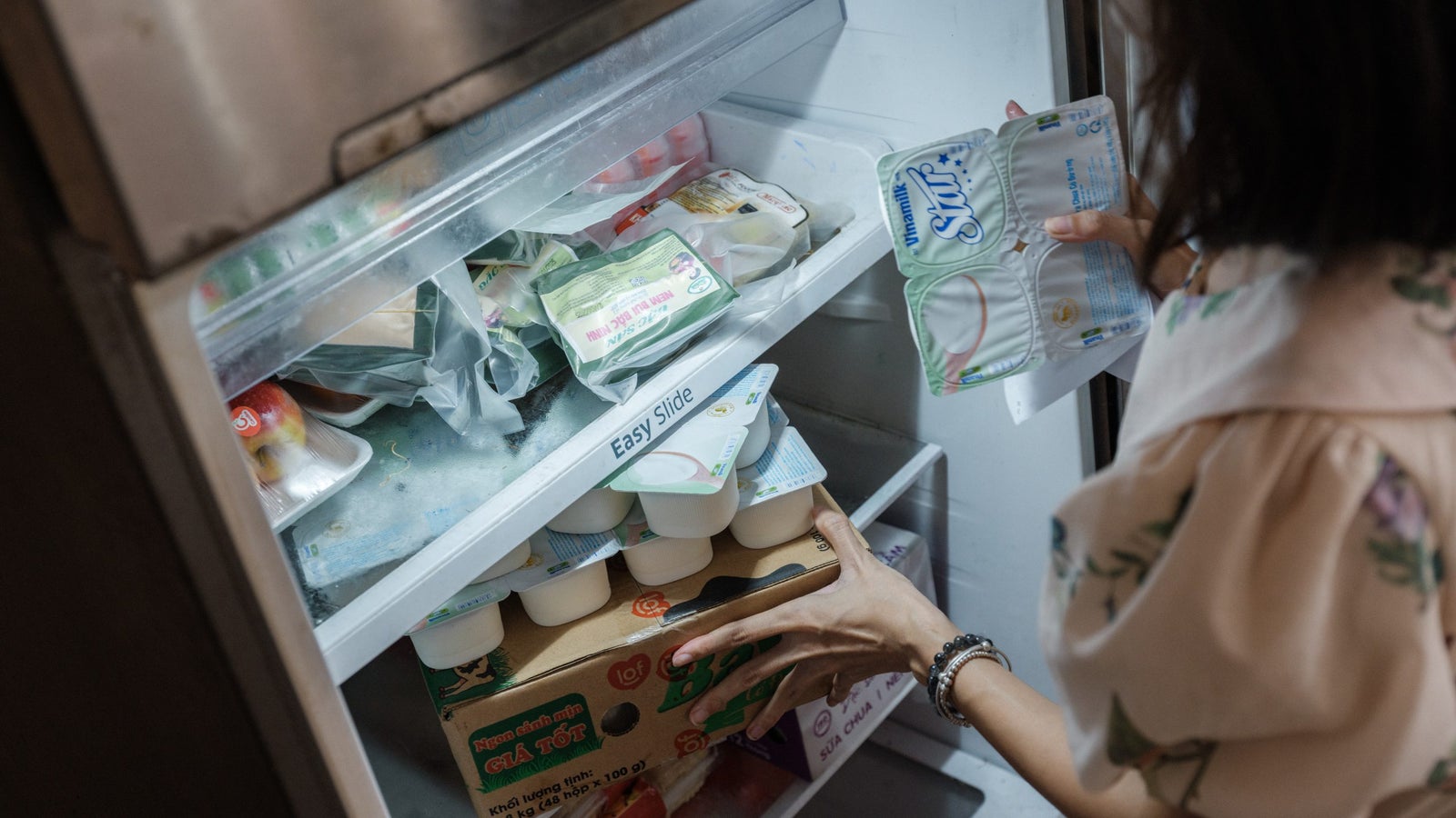 A person reaching into an open refrigerator, holding a package of yogurt while looking at various food items inside. The fridge contains containers of yogurt, eggs, and other food products on different shelves, showcasing a cluttered but organized arrangement.