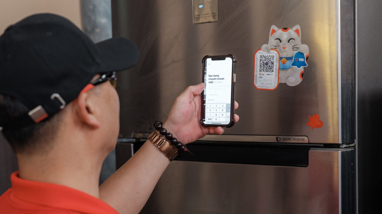 A person in a red shirt is using a smartphone to scan a QR code on a refrigerator adorned with a cartoon cat sticker, demonstrating a digital interaction.