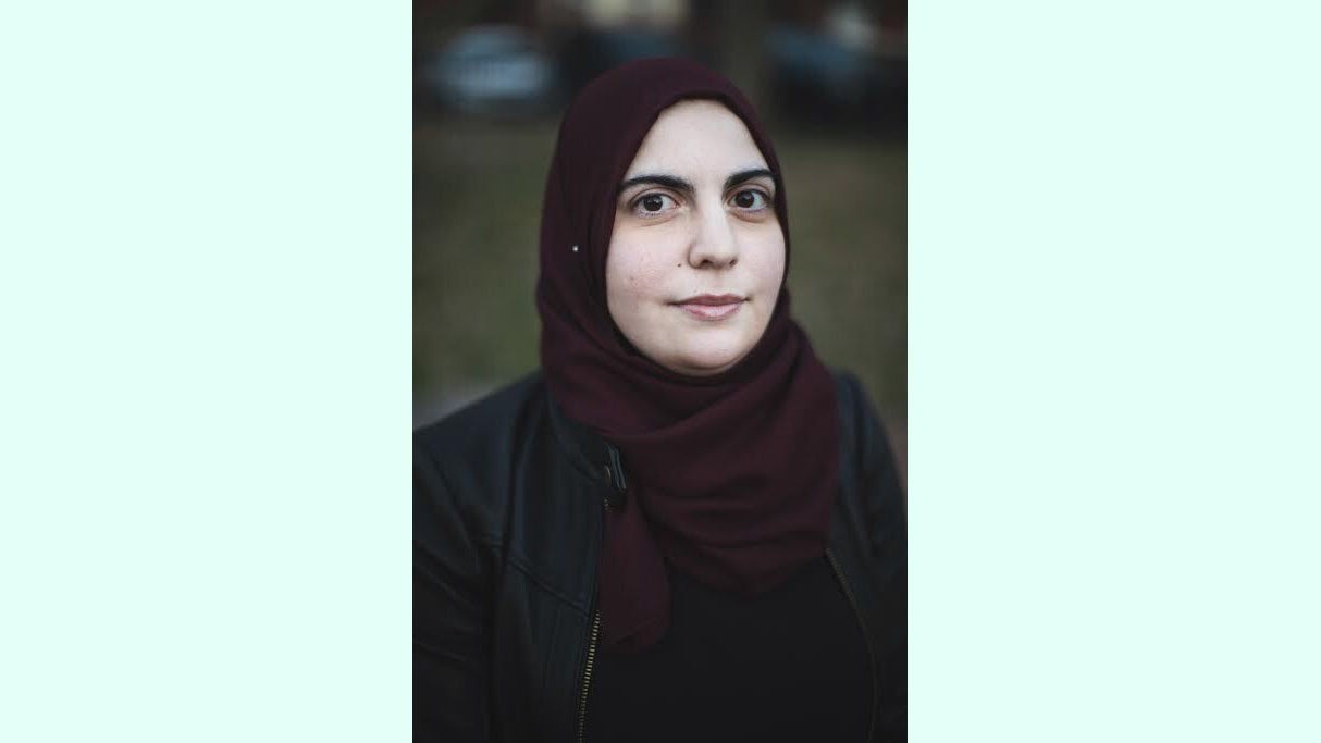 A portrait of a woman wearing a maroon hijab and a black jacket, looking directly at the camera with a neutral expression. The background is blurred, indicating an outdoor setting.