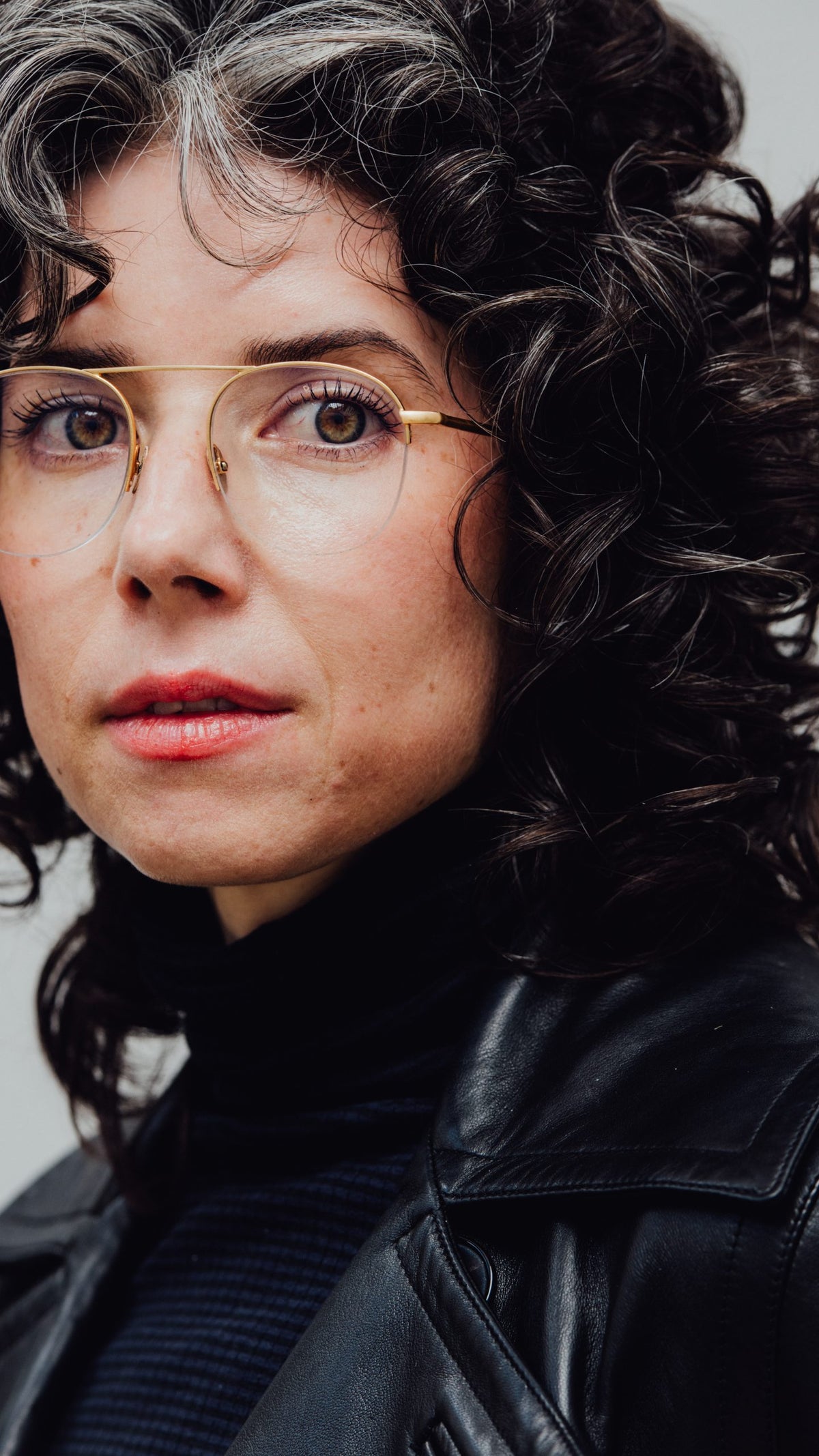 A close up portrait photograph of Signal President Meredith Whitaker standing in front of a plain wall, wearing a black leather jacket and black clothing.