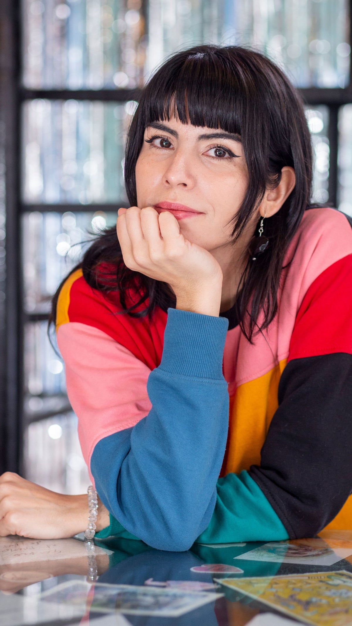 A woman with dark hair and bangs rests her chin on her hand, wearing a colorful, multi-paneled sweater. She is seated at a table covered with various colorful cards and papers, set against a blurred backdrop of shimmering decorations.