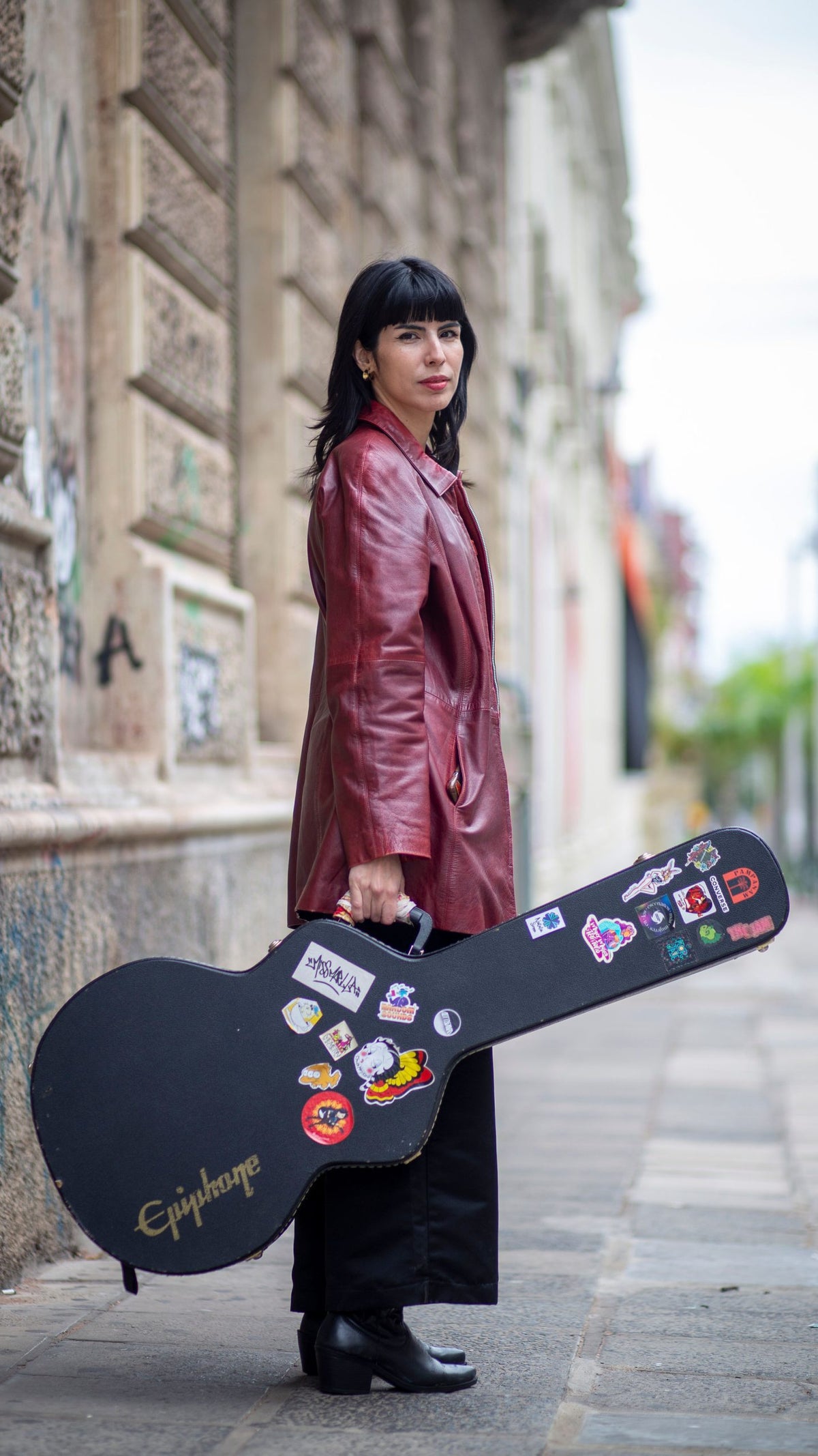 A woman with black hair stands on a street, wearing a red leather jacket and black pants, holding a guitar case covered in stickers, with a stone wall and blurred buildings in the background.