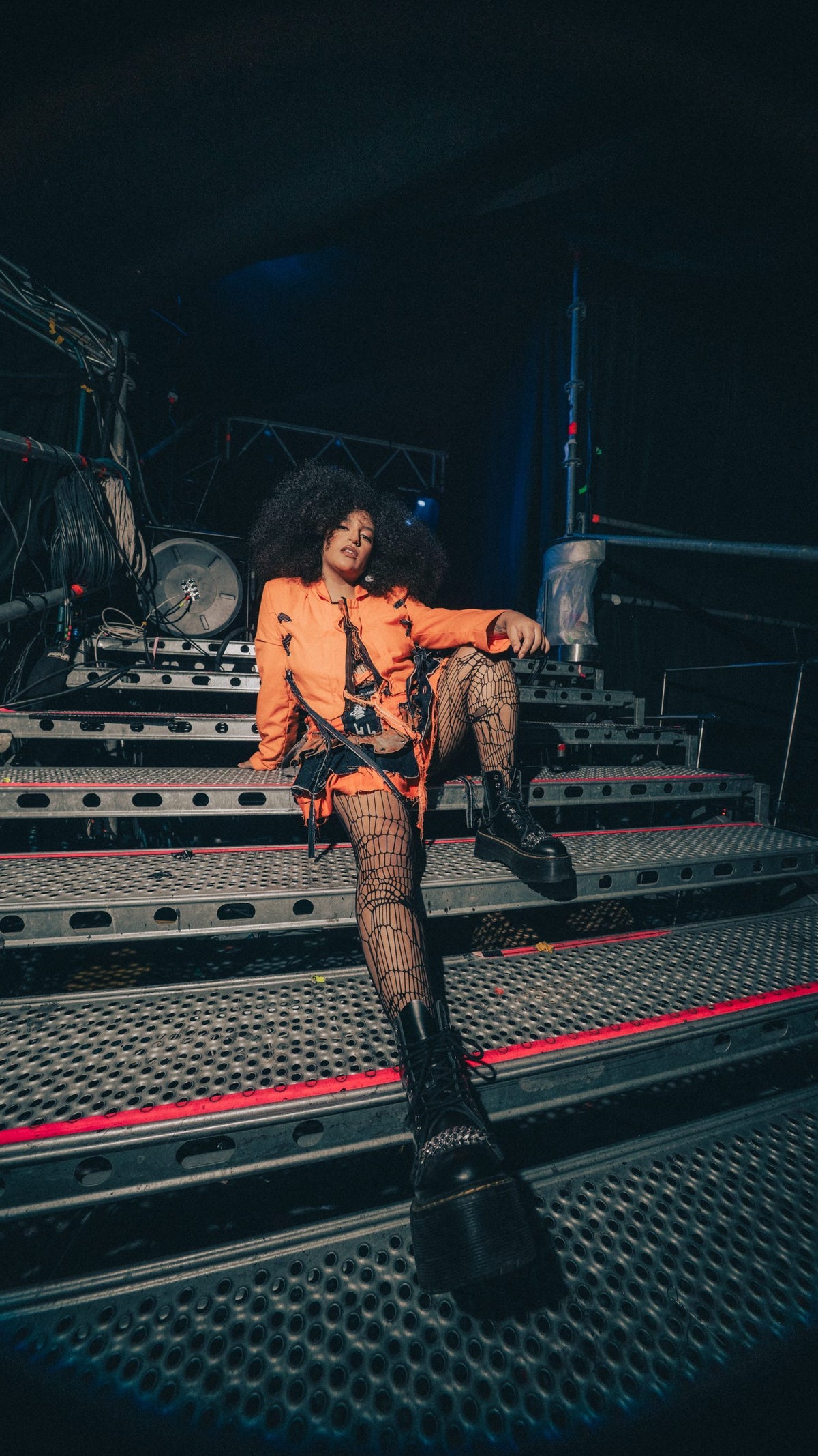 A person with large curly hair relaxes on a set of metal steps backstage, wearing an orange jacket over a patterned outfit and fishnet stockings, paired with chunky black boots, surrounded by stage equipment.