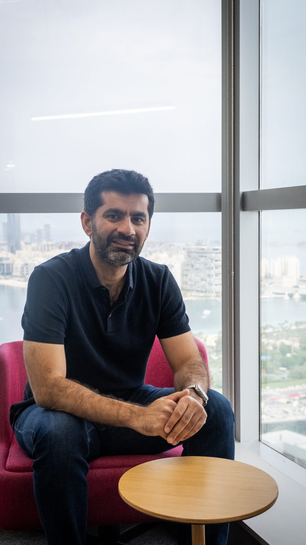 A man with a beard is seated on a pink chair next to a wooden side table, smiling and resting his hands on his knees. He is wearing a black polo shirt and blue jeans, with a city skyline and waterfront visible through the large window behind him.