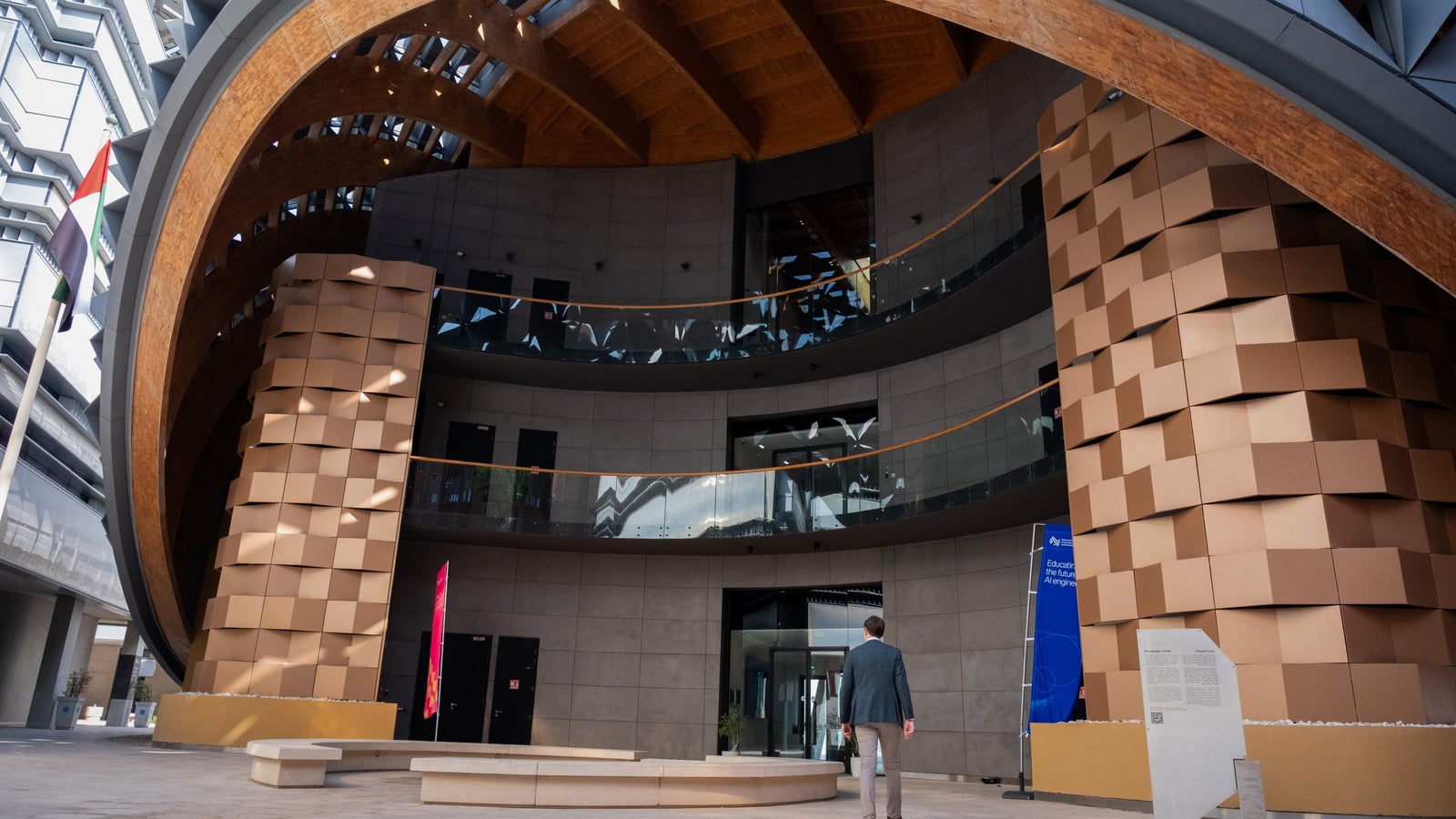 A person walks towards a modern architectural space featuring curved wooden ceilings, textured beige columns, and glass railings. There are banners in the background, and the floor is made of light-colored stone.