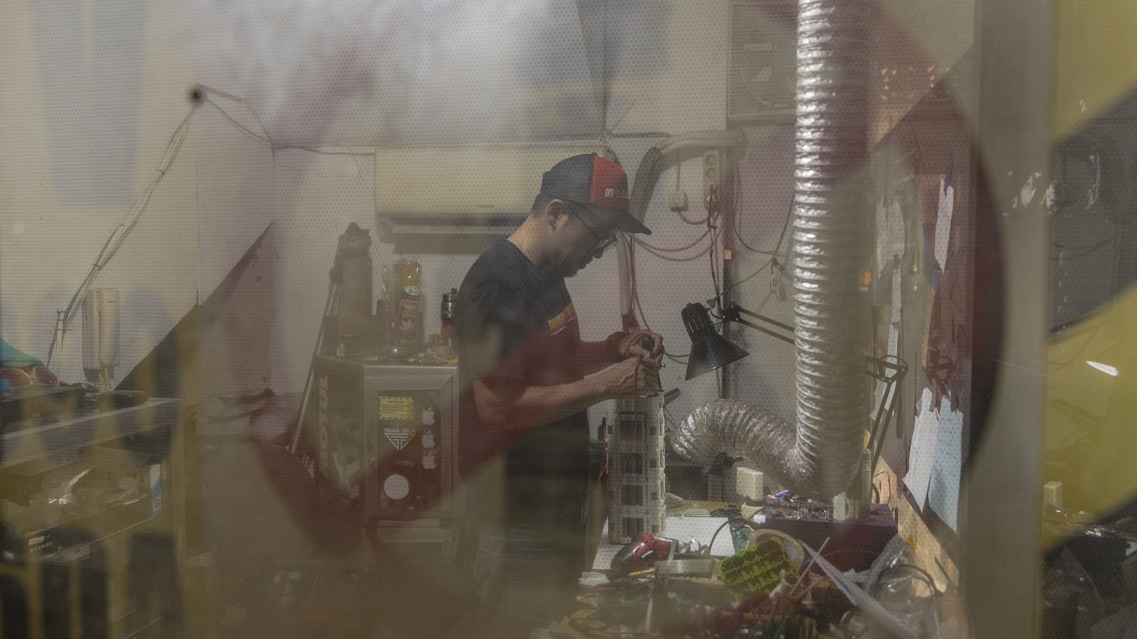 Dharmawan Kusna Handoyo works on repairing Wika Batt EV battery for motorbikes, seen through a clear door with a hazard tape and no entry sign visible.