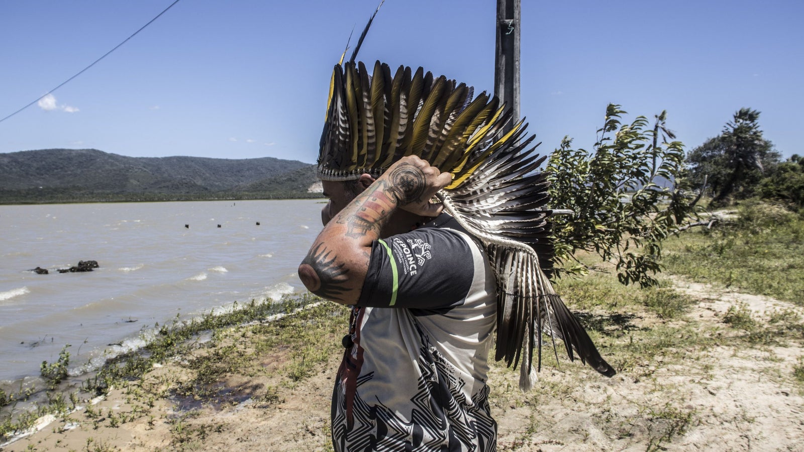 A person wearing a feathered headdress stands by a riverbank, looking out over the water. The landscape includes mountains in the background and greenery along the shore.