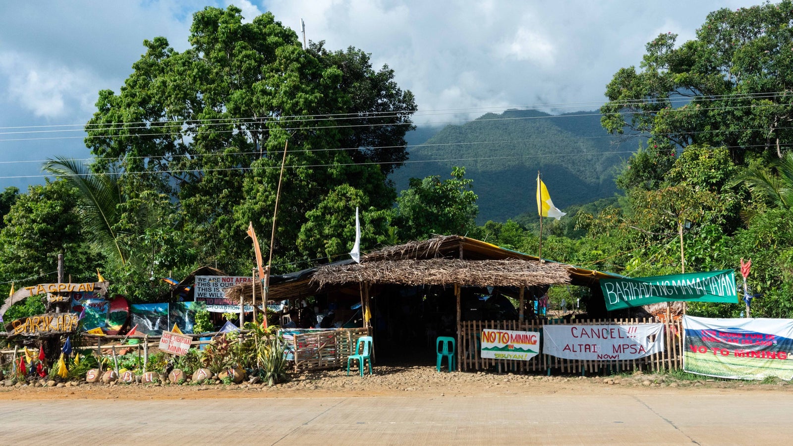 A barricade with protest signage seen from the street, blocking the access road to the mining area.