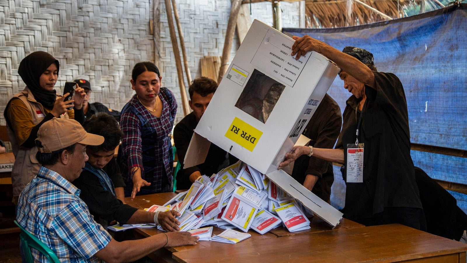 A photograph showing people standing around a table in a makeshift enclosure as a man pours out election materials from a box.