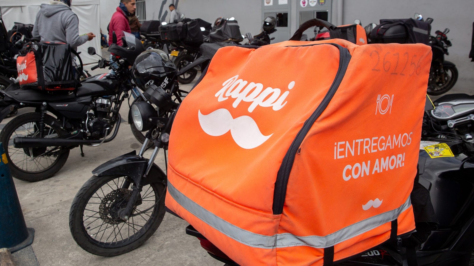 A photo showing a large orange Rappi delivery bag on the back of a motorbike, parked alongside other bikes.