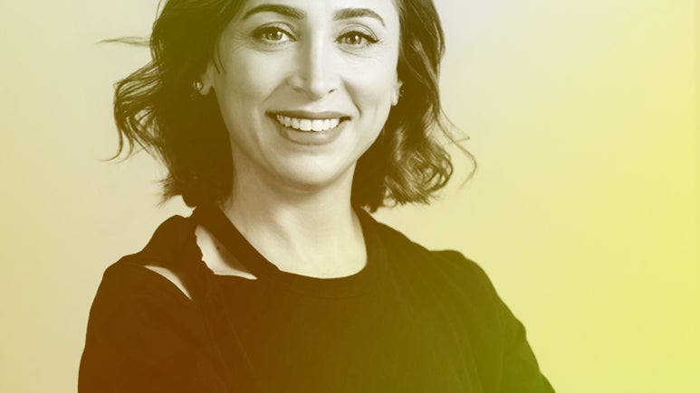 A woman with shoulder-length wavy hair smiles confidently, wearing a black shirt against a softly colored background.