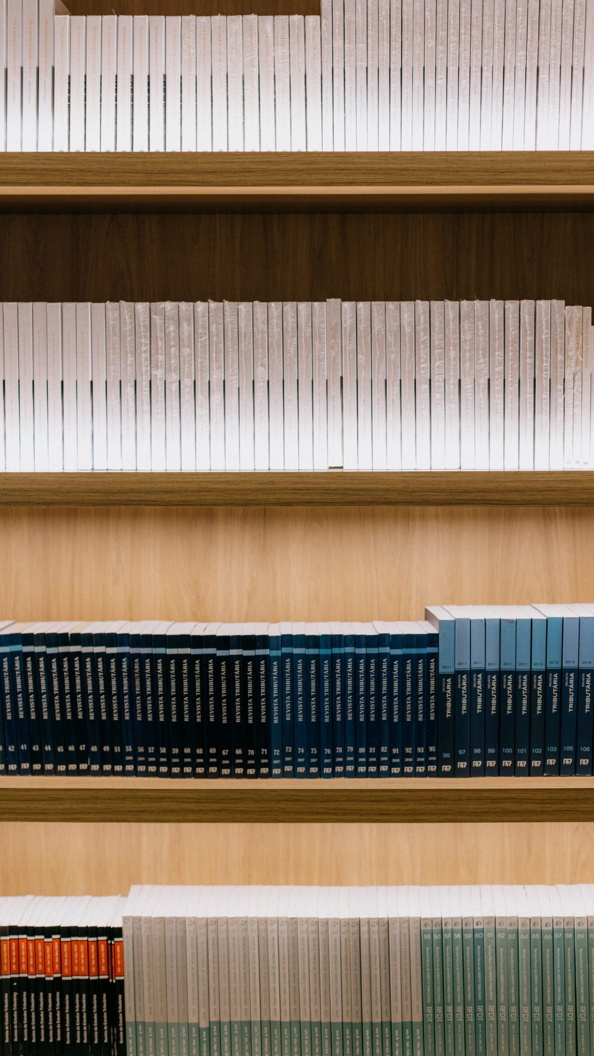 A wooden bookshelf displaying multiple rows of uniform books, with the top shelf filled with white books, the middle shelf featuring dark blue and turquoise books, and the bottom shelf containing a mix of light blue and white books, organized neatly.