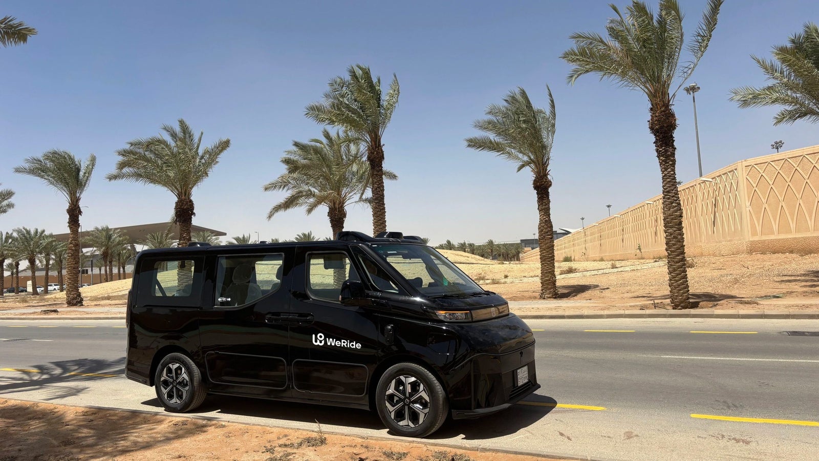 A black autonomous vehicle with the logo 'WeRide' parked on the roadside, surrounded by palm trees and a clear blue sky in a desert-like environment.
