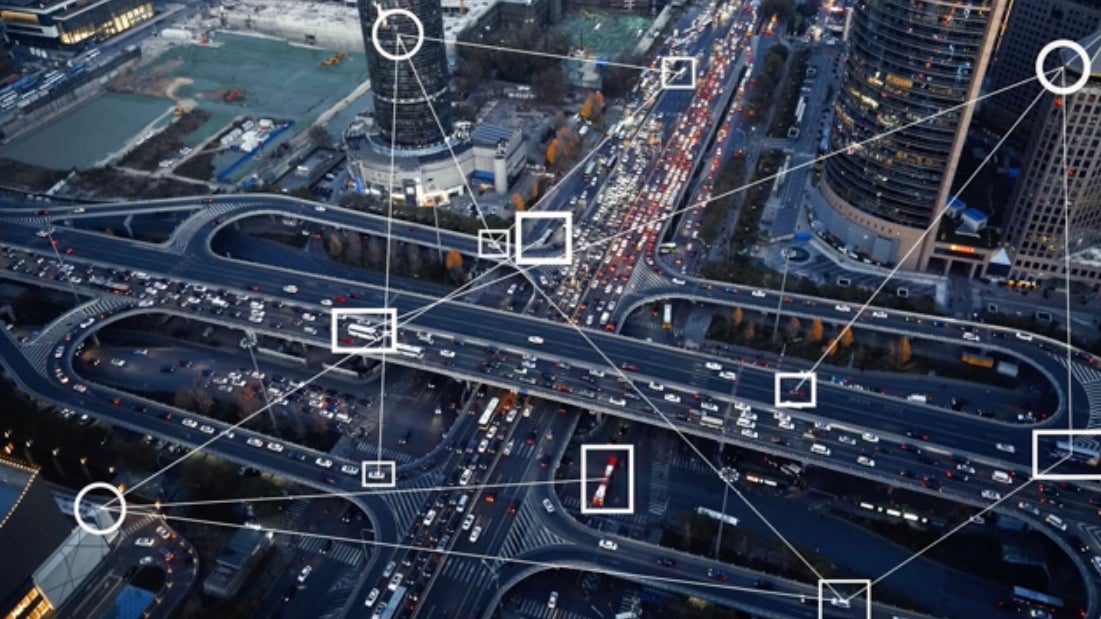 Aerial view of a busy urban intersection with vehicles on multiple roadways, overlaid with digital tracking outlines, suggesting a network analysis or smart city infrastructure.