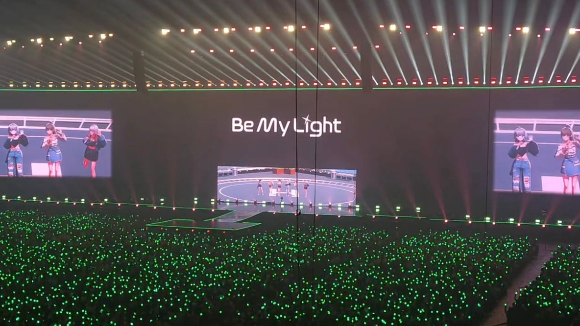A large concert venue filled with fans holding green light sticks, with a prominent stage displaying the words "Be My Light" and screens showing performers engaging in a dance or performance.