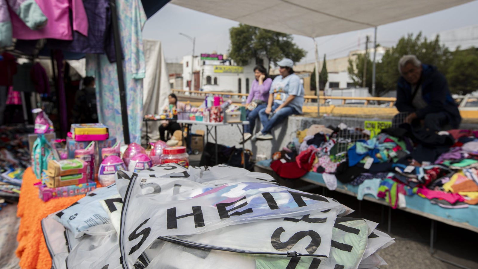A bustling outdoor market scene featuring a variety of clothes and colorful products scattered on tables. In the foreground, transparent bags with printed labels are stacked, while vendors sit on the edge of a concrete surface, engaging with customers. Brightly colored garments and other items are displayed on nearby tables.