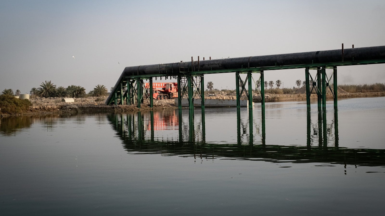 A large oil pipeline supported by green pillars stretches over calm water, reflecting the structure and nearby palm trees.