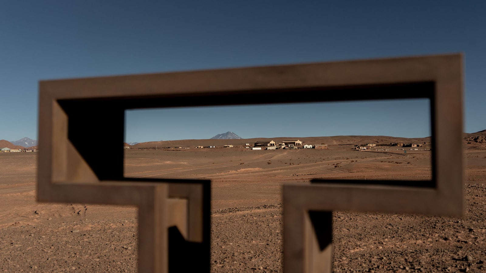 A metal frame in the foreground outlines a view of a distant mountain and a sparse landscape dotted with a few buildings and scattered vegetation, under a clear blue sky.