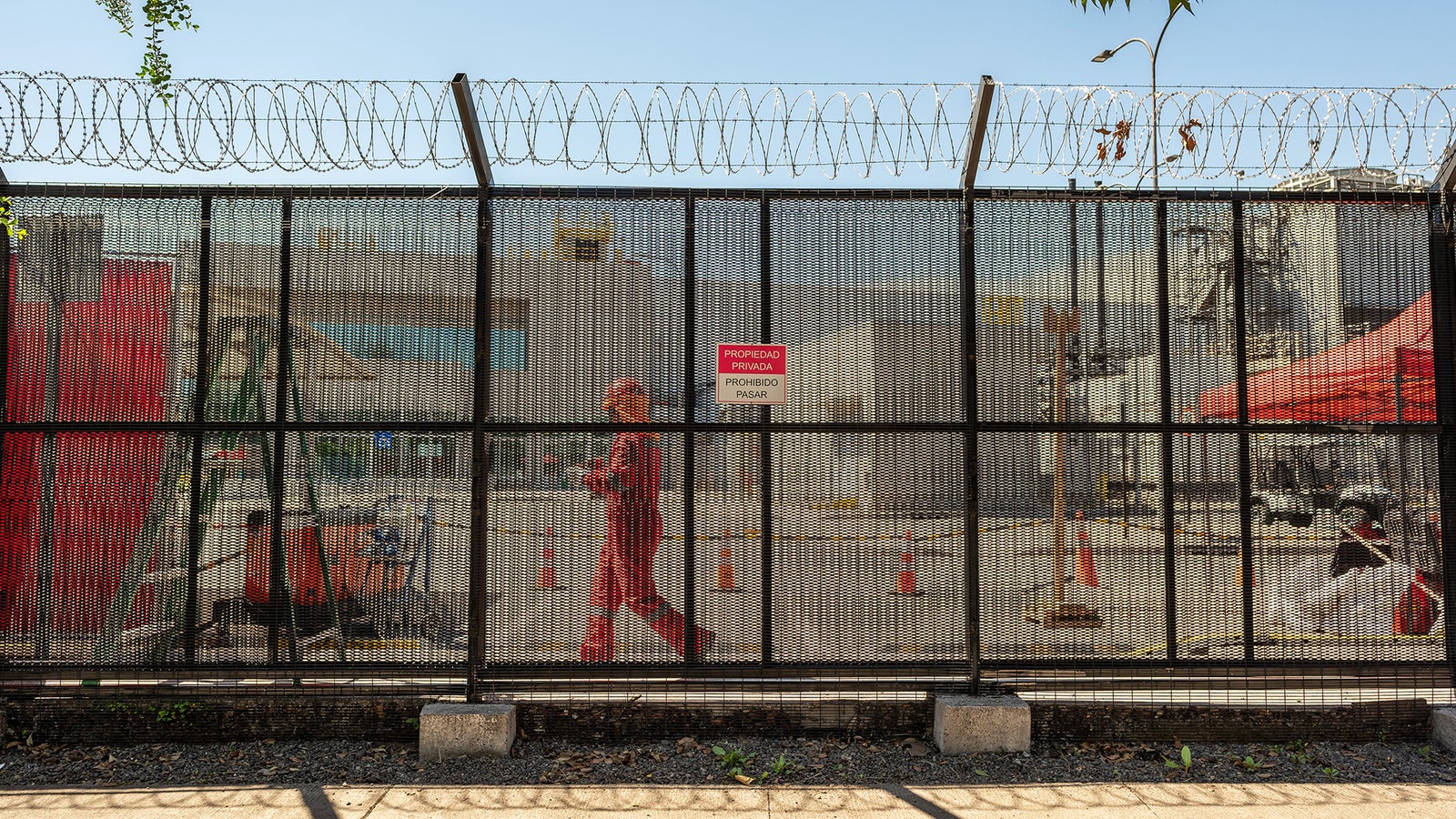 A mesh fence topped with barbed wire, containing a sign prohibiting entry, with a worker in an orange suit walking past.