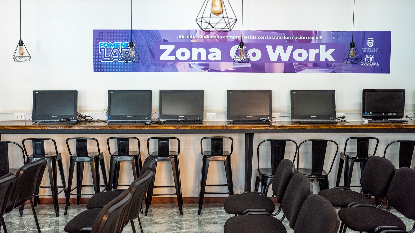 A coworking space with multiple desktop computers on a long table, black stools, and pendant lights above. A banner reads 'Zona Co Work'.