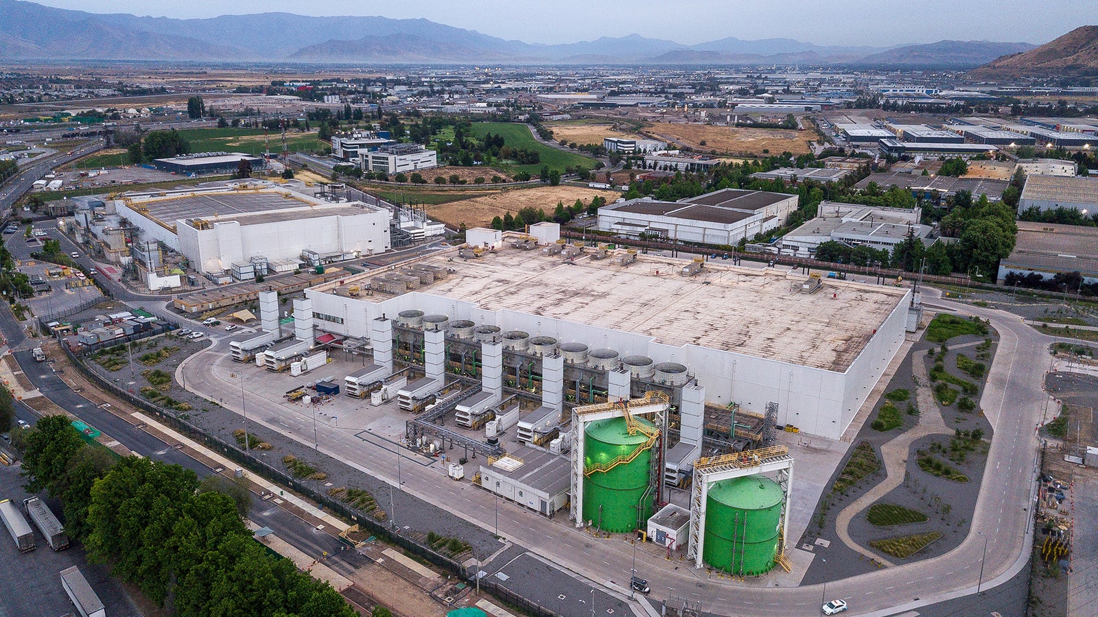 Aerial view of an industrial facility with large storage tanks and trucks, surrounded by green fields and mountains in the background.