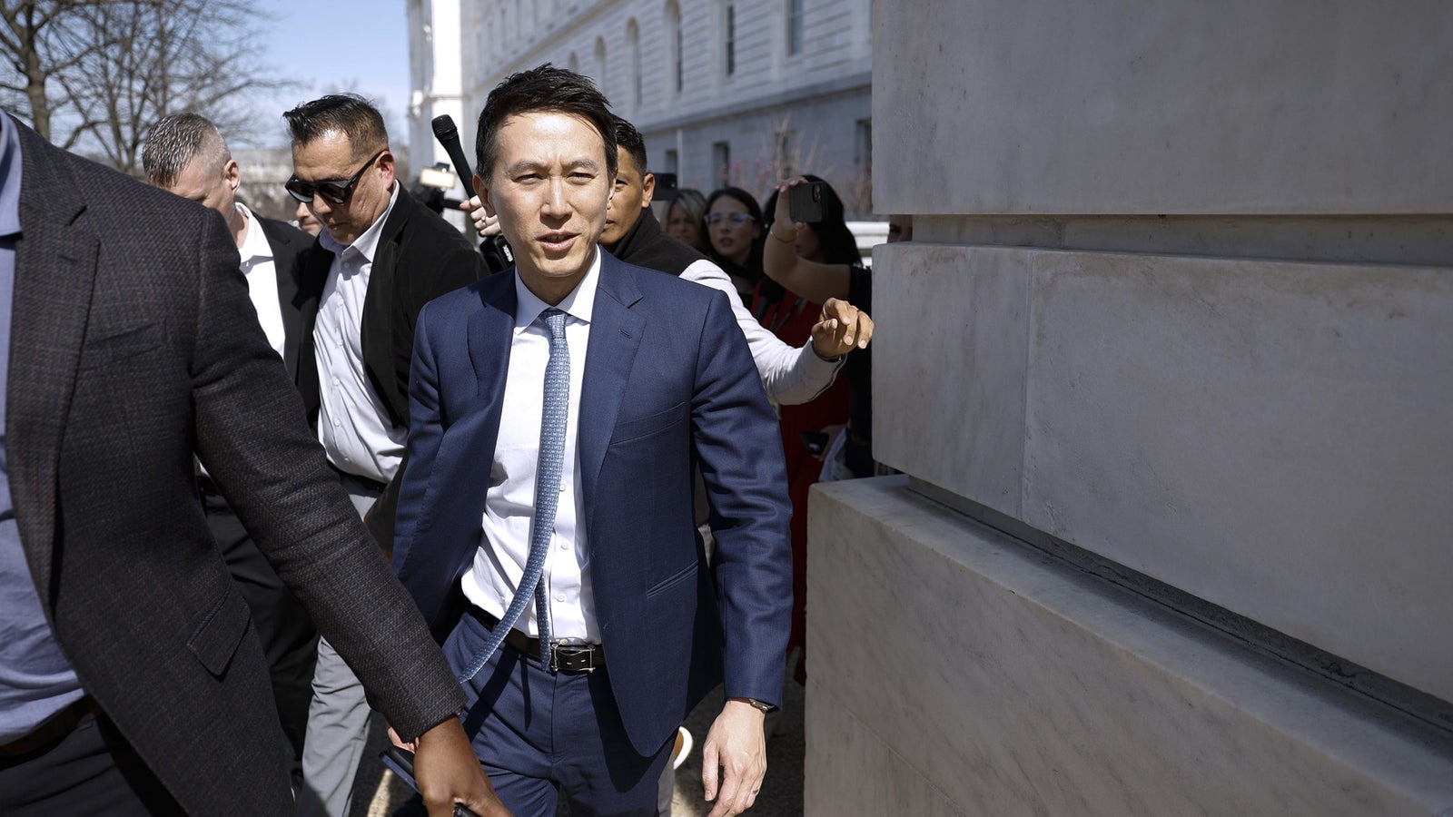 A man in a blue suit walks amidst a crowd, flanked by security, in front of a government building.