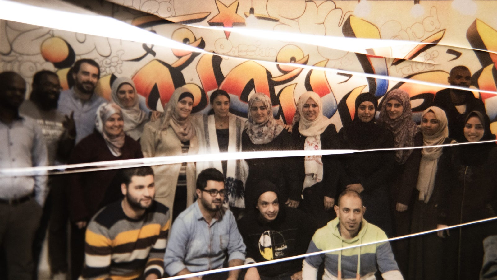 A diverse group of individuals posing together in front of a colorful graffiti wall; the group includes men and women, with several wearing headscarves, and they are arranged in two rows, some standing and others seated, conveying a sense of camaraderie and community.