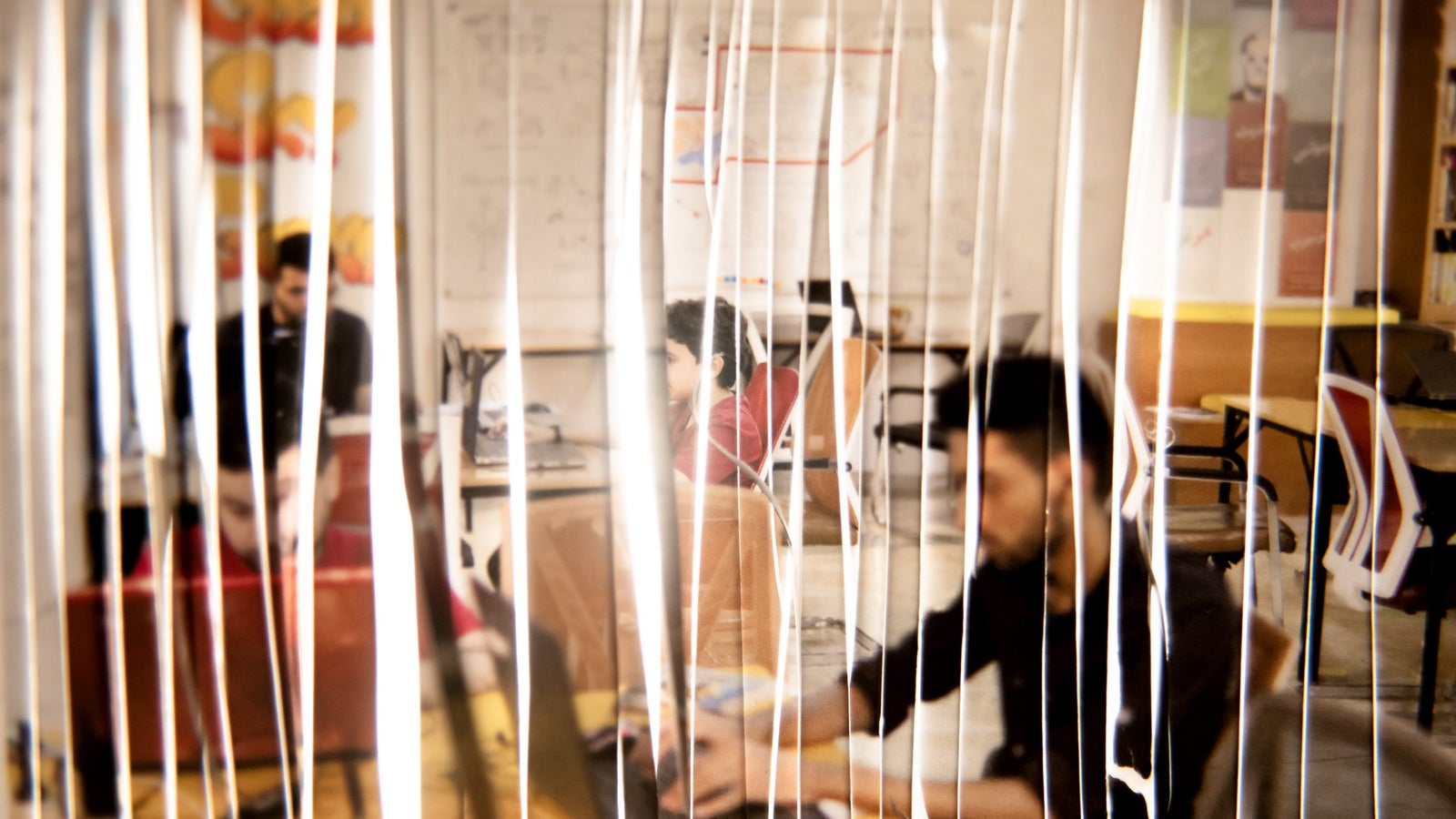 A blurred interior of a workspace with thin vertical lines in the foreground, showing several individuals engaged in work at their desks. The background includes a wall with colorful posters and a whiteboard filled with drawings.