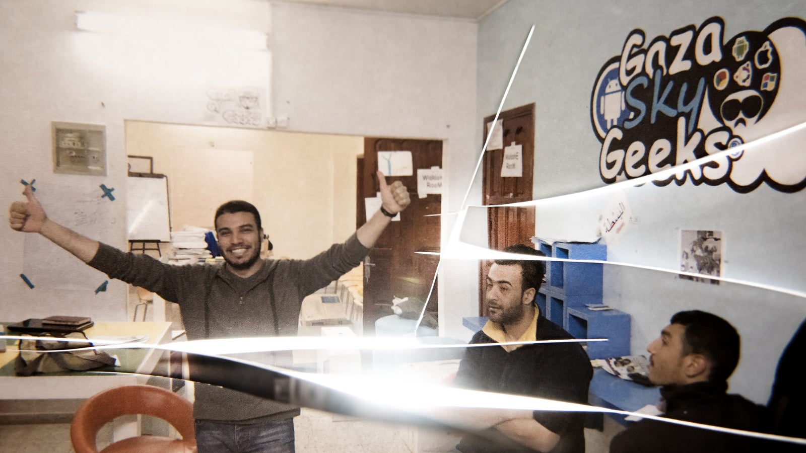 A group of young men in a casual workspace, with one man giving a thumbs-up and smiling, while two others sit attentively. The background features a logo for 'Gaza Sky Geeks' and blue shelving. The lighting creates streaks across the image.