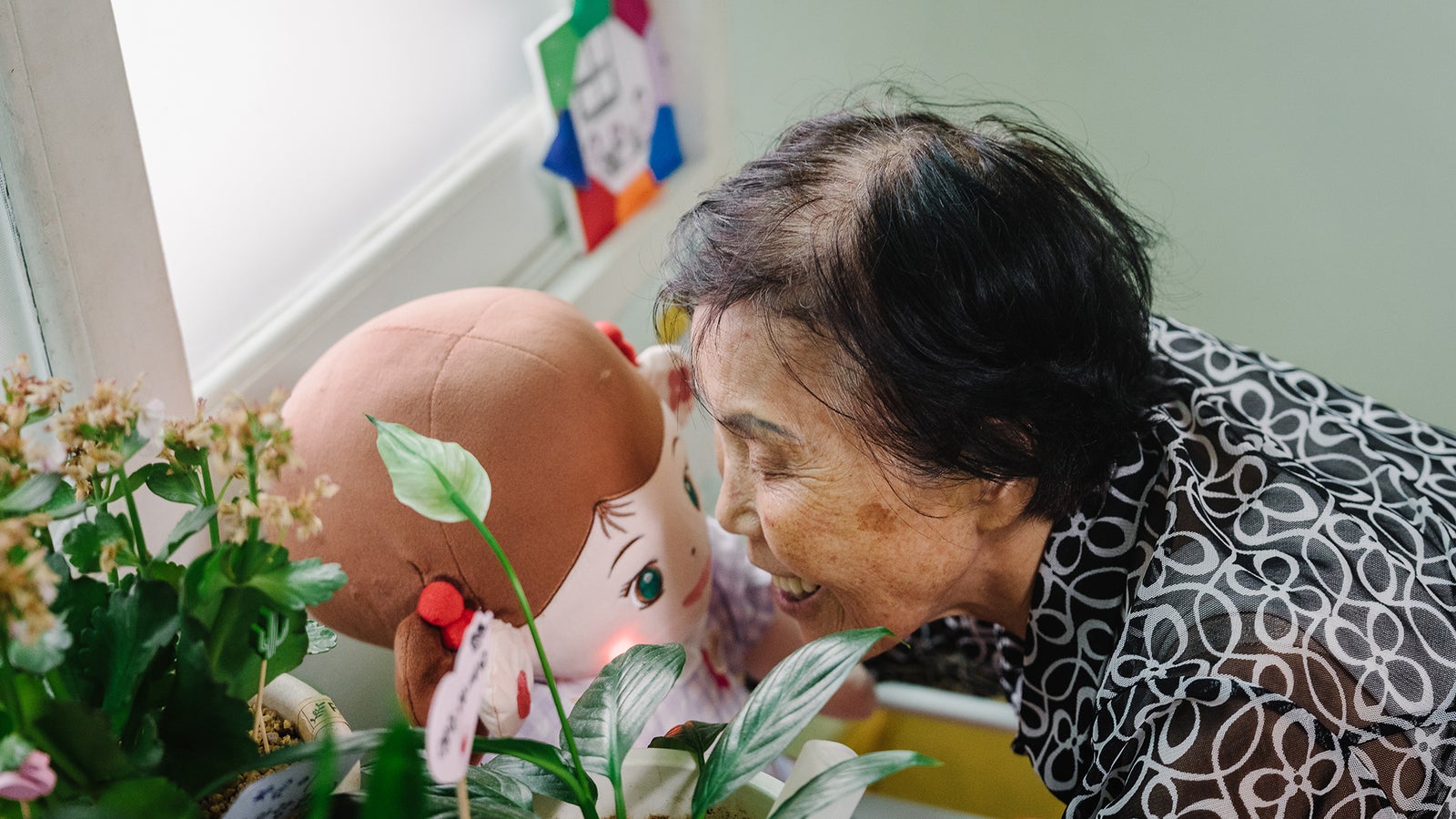 An elderly woman with short black hair smiles as she leans in close to a plush doll resembling a girl with brown hair, surrounded by potted plants and colorful decorations on a window sill.
