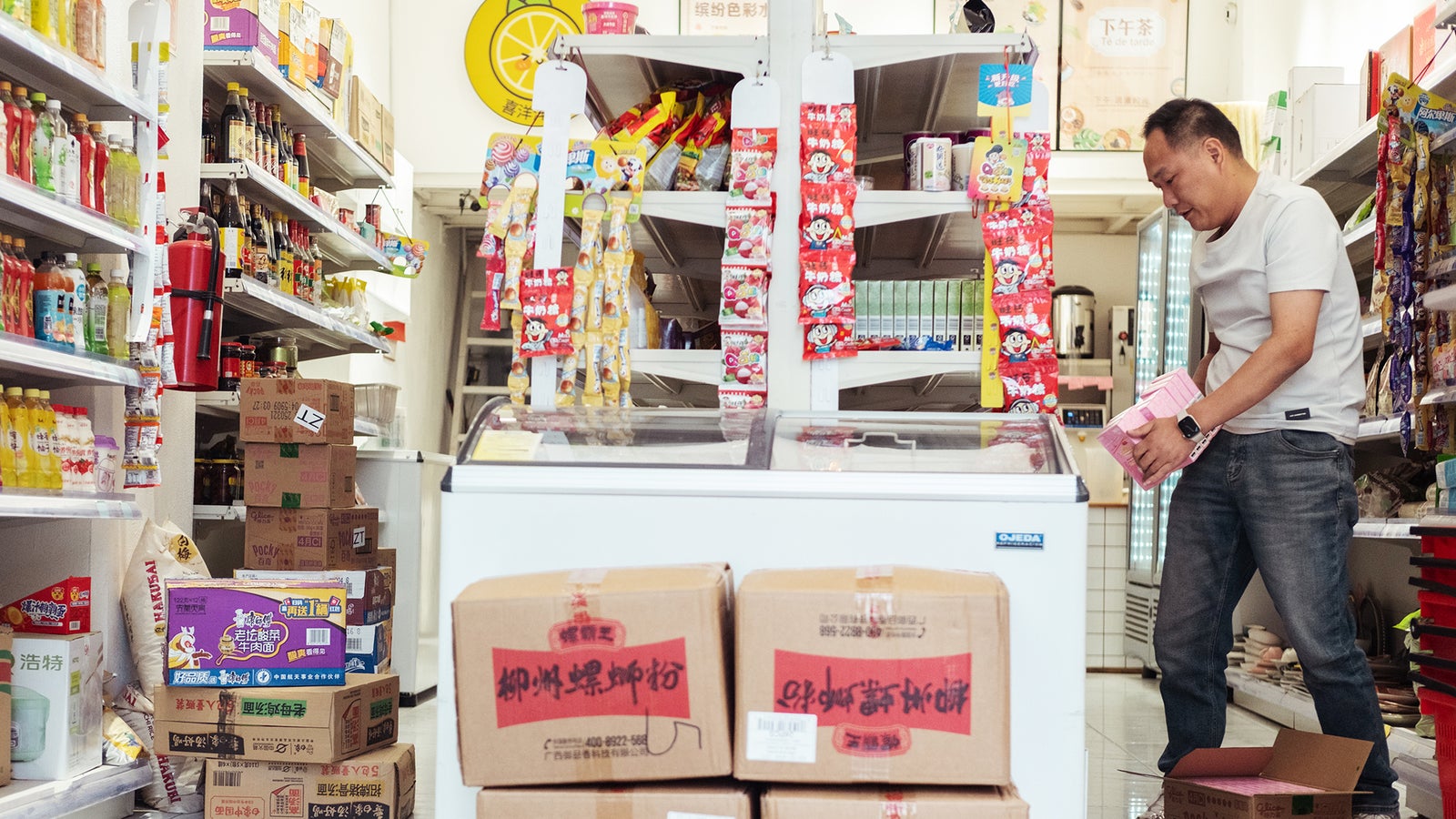 A man in a white shirt is stocking shelves in a small convenience store, surrounded by colorful products and cardboard boxes.