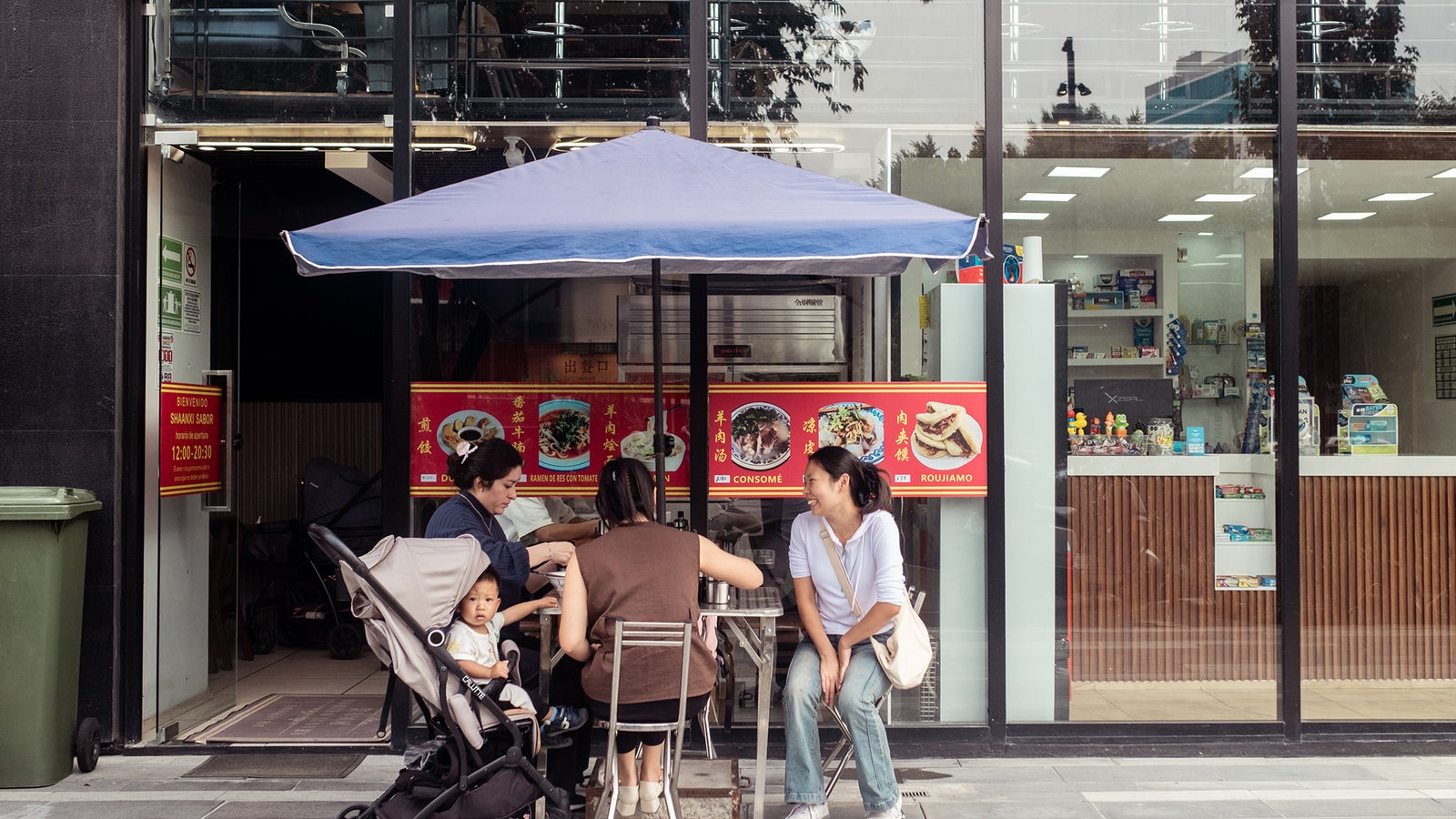 A family enjoys a meal at an outdoor table under a blue umbrella in front of a restaurant with a display of food images.