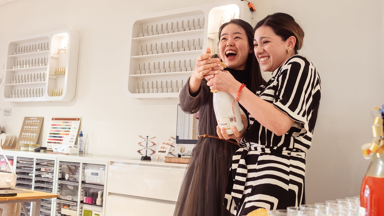 Two women joyfully holding a bottle of sparkling drink in a nail salon with shelves of nail products behind them.