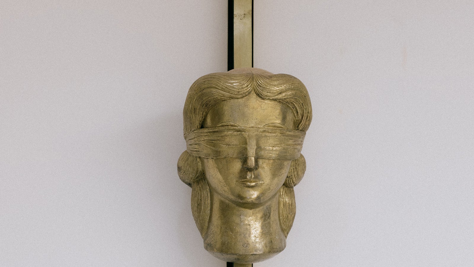 A gold-toned sculpture of a blindfolded female figure's head, featuring wavy hair and a smooth face, mounted on a vertical pole against a plain white background.