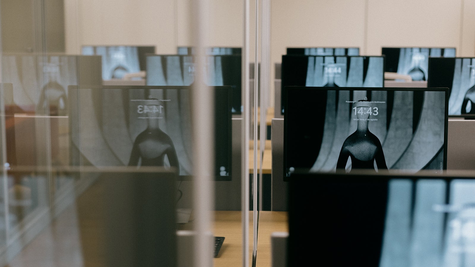 An office space with multiple computer monitors displaying a similar image of a torso, arranged in rows and divided by glass panels, with the time visibly shown on the screens.