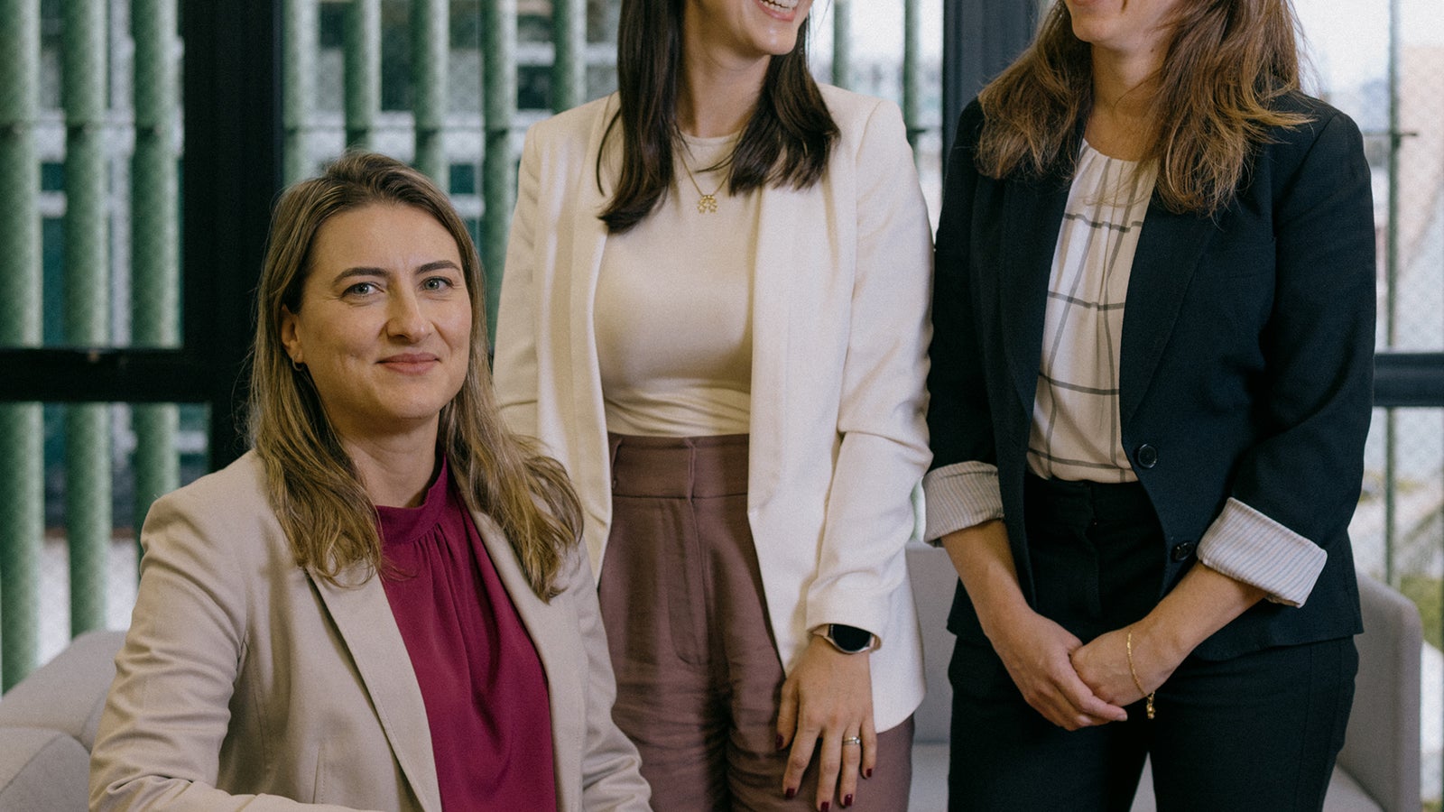 Three women are standing together in a modern office setting. The woman in front is seated, wearing a light-colored suit and a burgundy top, while the other two women are standing, smiling, in white blazers and dark business attire. The background features large windows and a green interior design.