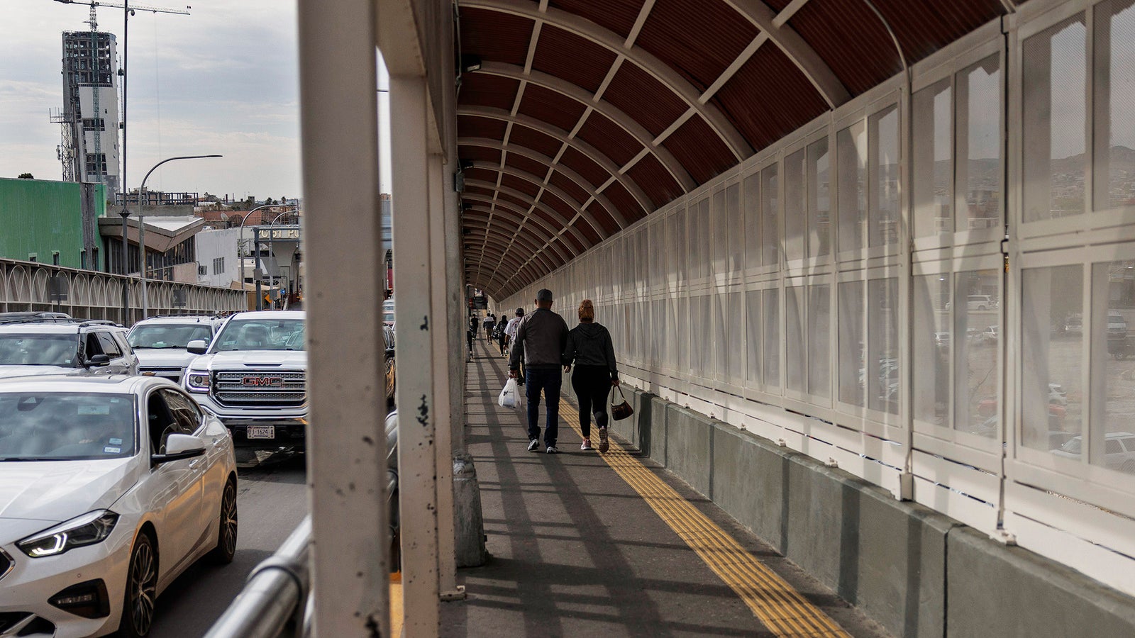 Two people walk side by side in a covered walkway, with cars visible in the background and urban structures lining the street.