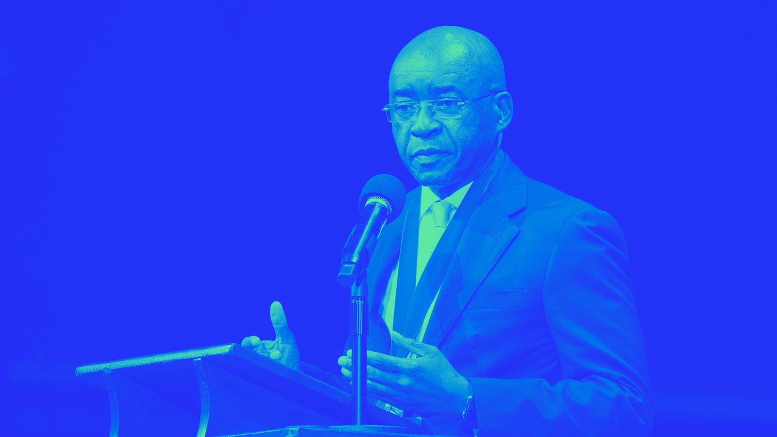 A man in a blue suit and tie stands at a podium, gesturing with his hands while speaking into a microphone.