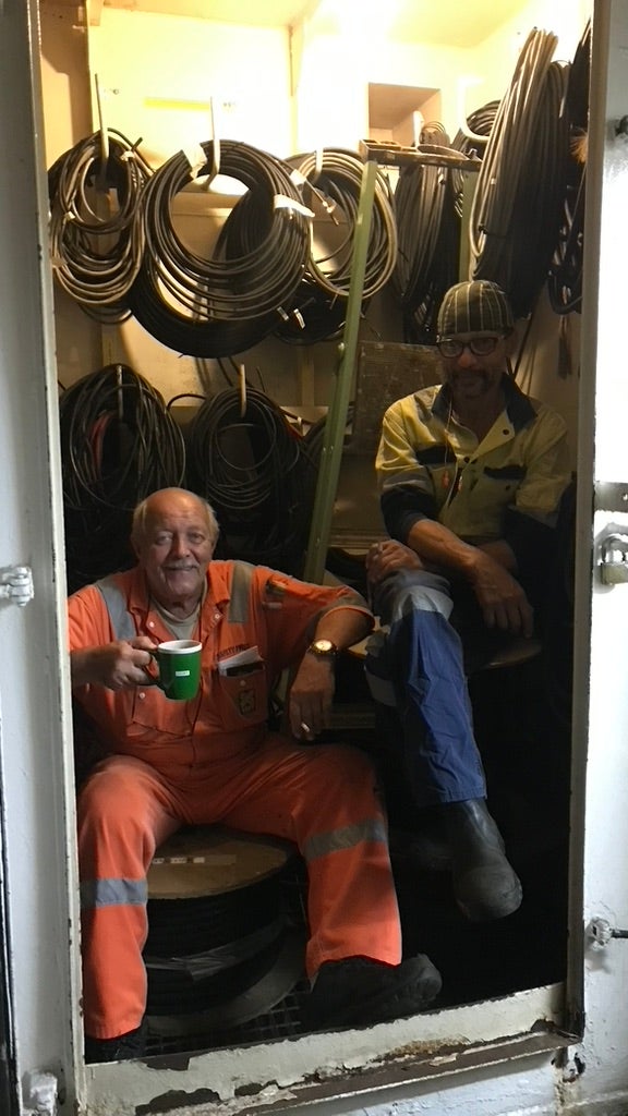Two men seated inside a cramped space filled with coiled cables, the man on the left wearing an orange jumpsuit cups a green mug, while the other, in a yellow shirt and glasses, sits beside him.