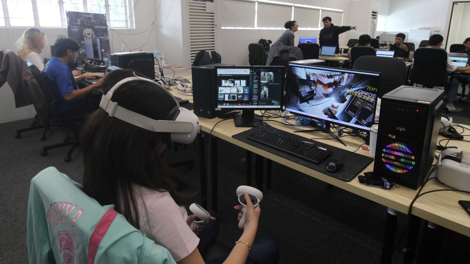 A group of people working in a computer lab, with one person wearing a virtual reality headset and holding controllers, while multiple monitors display various screens. The workspace features several desktop computers and cables, and other individuals are engaged in different tasks in the background.